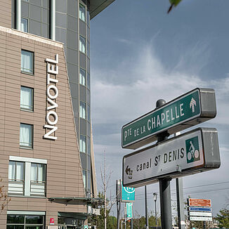 Street signs in Paris with the facade of the Novotel Hotel in the background.