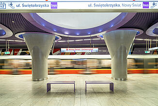 Passing train in a metro station in Warsaw with ceramic tiles in the waiting area and purple-coloured elements on the walls and ceilings.