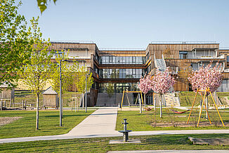 Exterior view with wooden façade, a playground, green areas and stairs leading to the terraces of the educational campus in Vienna.