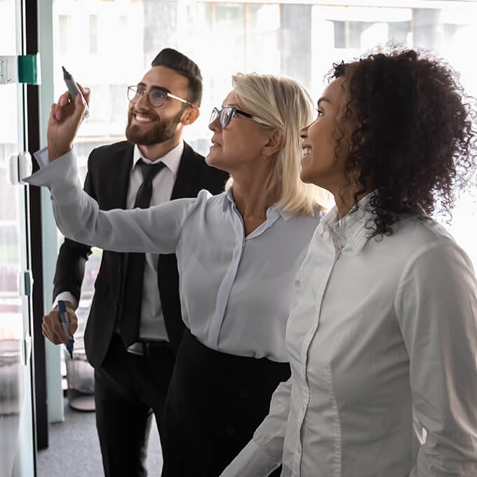 Two women and a man are standing in front of a whiteboard in an office.